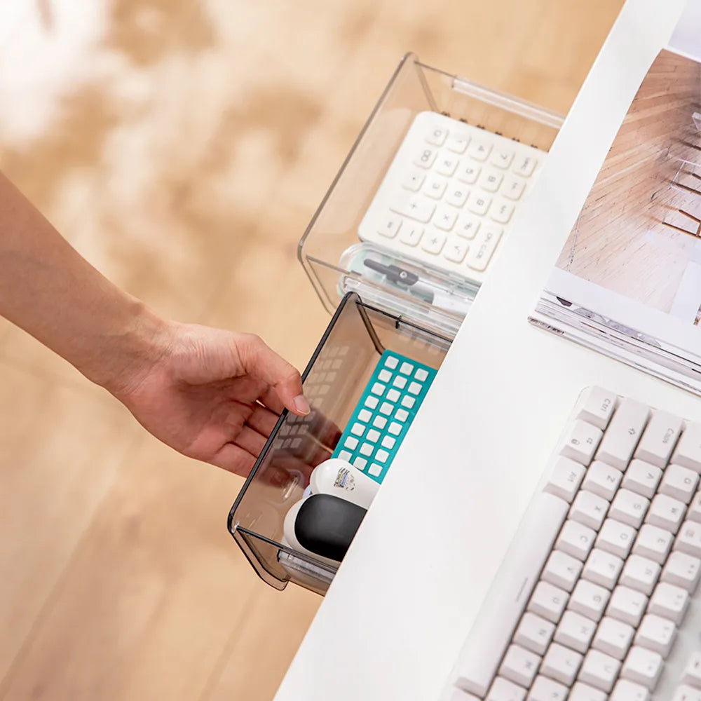 Hand pulling out a clear under-desk drawer showcasing easy access to neatly stored stationery items, next to a white mechanical keyboard on a desk.