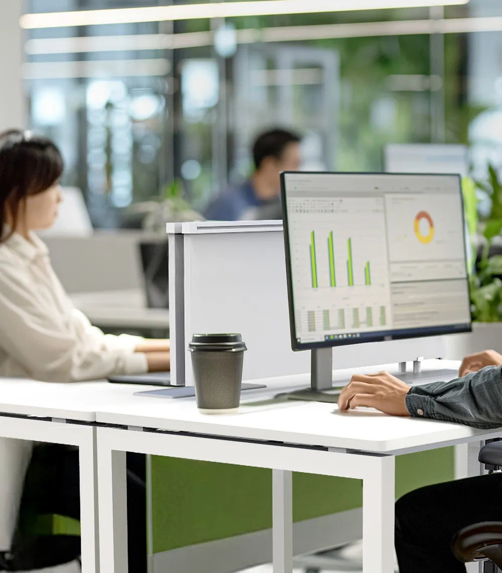 Two professionals working in a modern office equipped with ergonomic furniture. The focus is on a man sitting in a comfortable ergonomic chair, facing dual monitors displaying charts and data. The text overlay reads, "Customize. Optimize. Maximize Productivity."