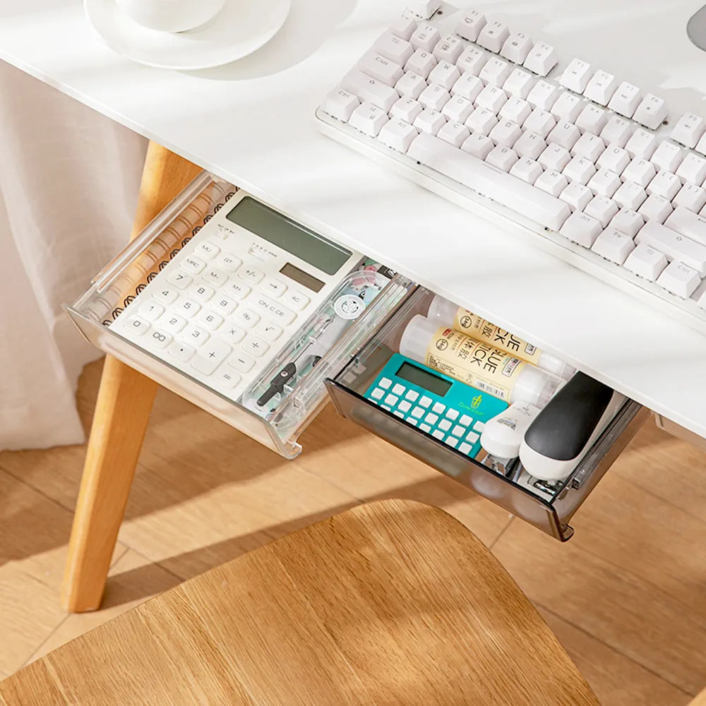 Transparent under-desk storage drawer filled with office supplies, mounted beneath a white desk with a wooden chair in the background, enhancing a neat workspace.