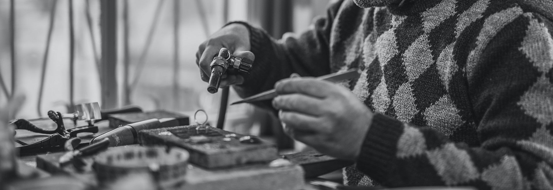 Monochrome image of a person's hands crafting or repairing items on a workbench, illustrating the detailed work, with the Flujo logo and navigation menu at the top.