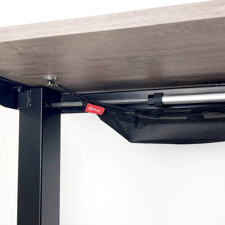 Under-desk cable management net with cables neatly tucked away, featuring the Flujo logo on a black strap, attached to a wooden desk with a metal frame.